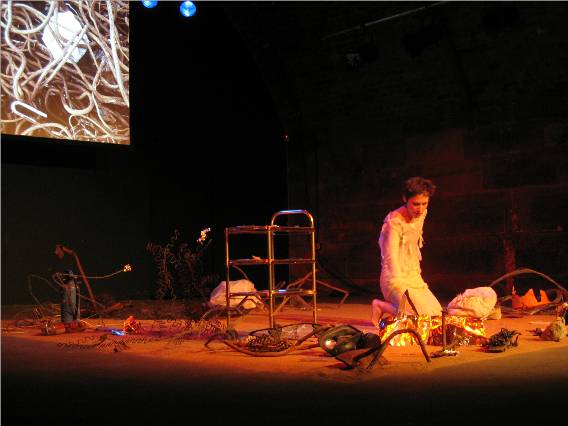 A woman kneels on a stage in reddish

 light surrounded by variouis objects, 

with an inset showing a tangle of wires in the top left corner.